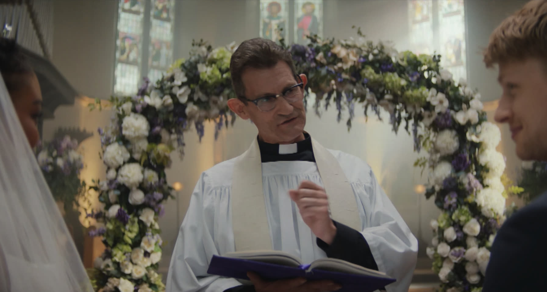 A vicar mid-ceremony under a floral arch, a bride and groom either side